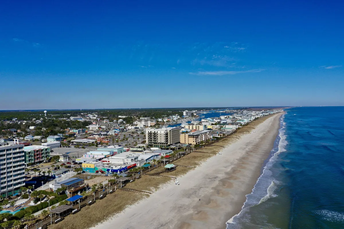A relaxed view of the beachfront with coastal buildings, rolling ocean waves, and the inviting proximity to waterfront dining options. The scene hints at easy access to the Carolina Beach Boardwalk, bustling with activities, eateries, and nightlife.