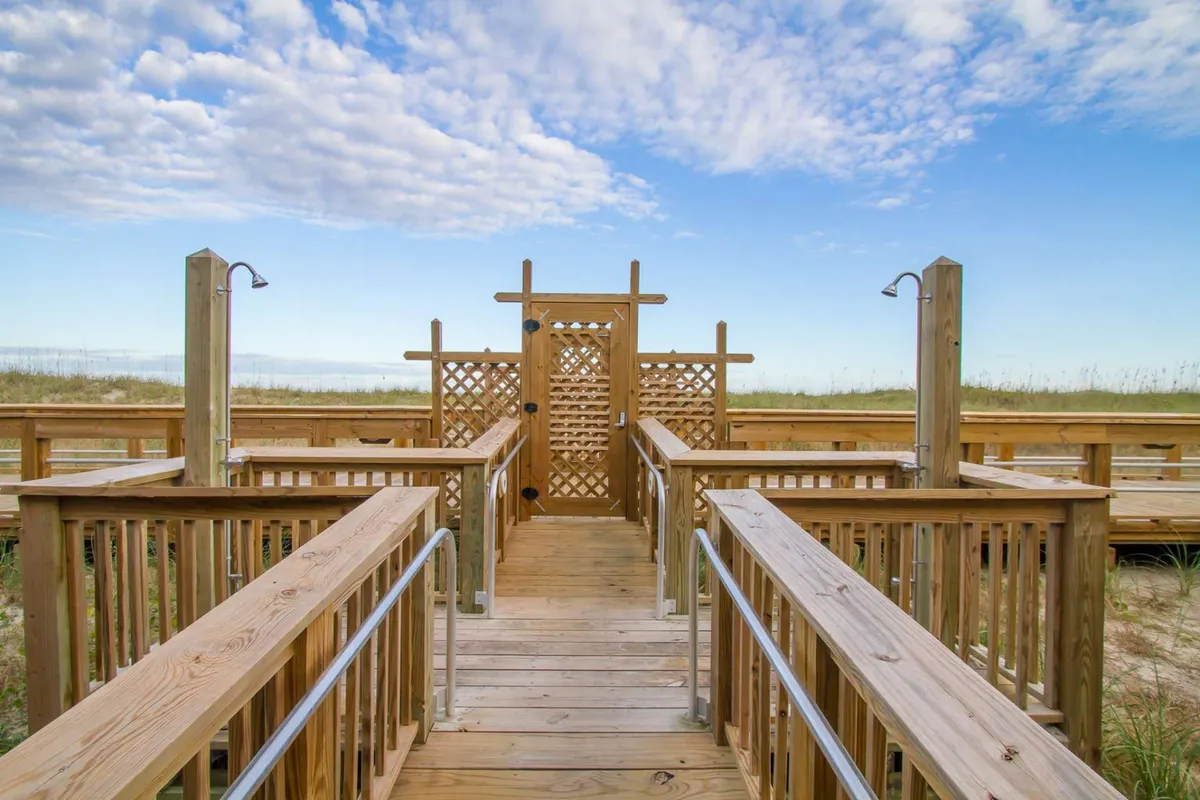 A tranquil wooden boardwalk with a decorative lattice gate under a blue sky. The boardwalk leads towards the ocean, visually evoking seamless private access to the beach.