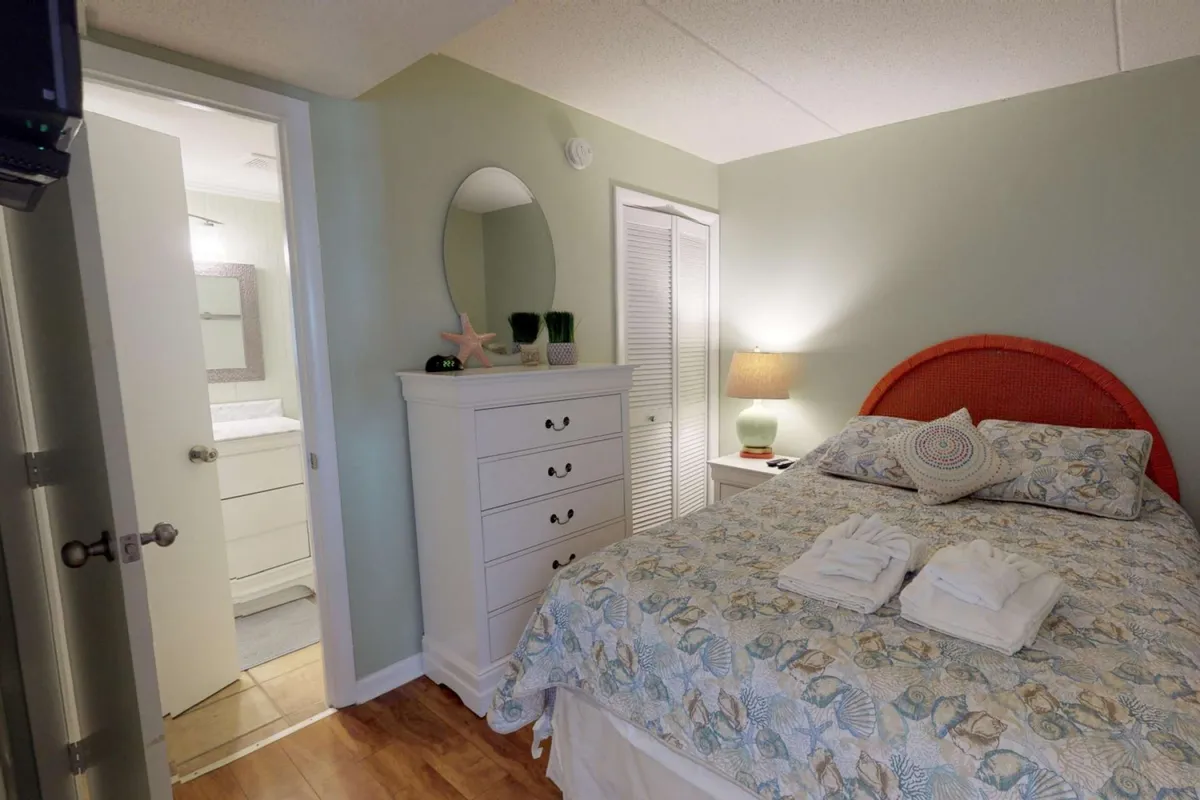 A cozy bedroom angle with a bed adorned in decorative pillows, a nightstand lamp, a dressing mirror, and a dresser with charming seashell decor. The atmosphere is serene and welcoming, perfect for unpacking and settling in.