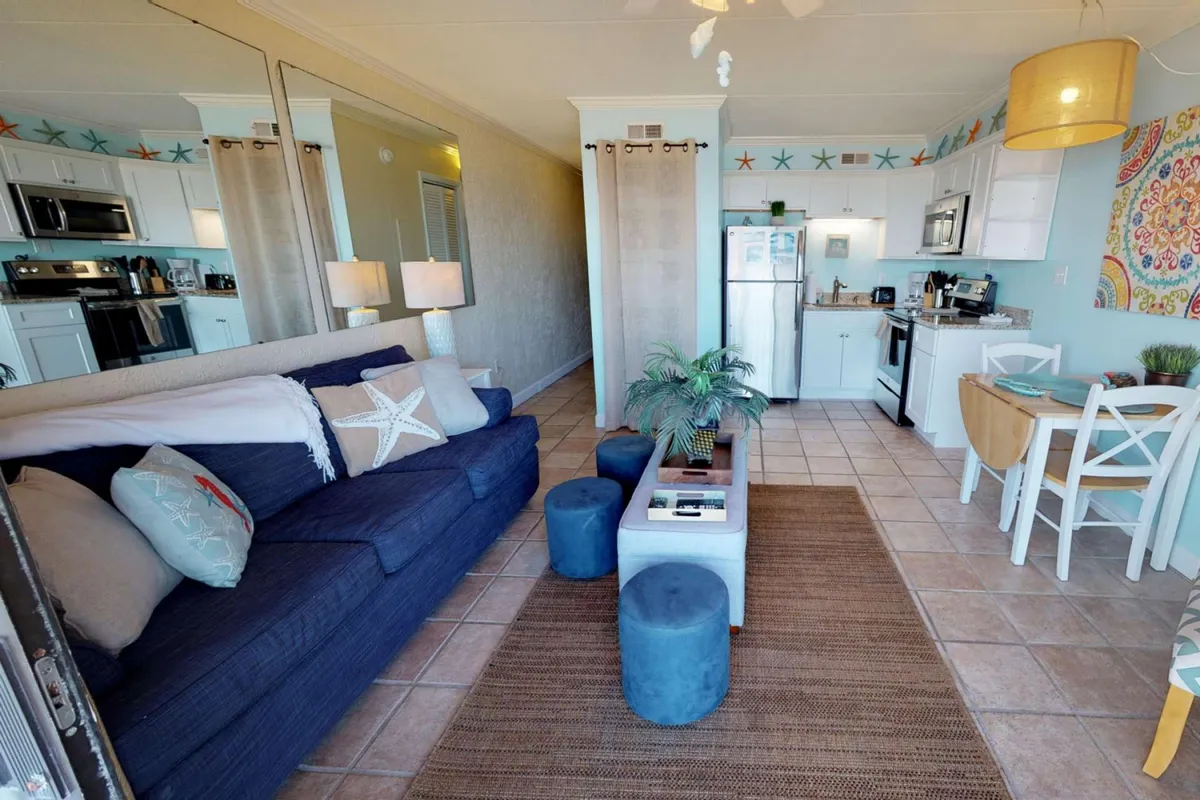 A wide-angle view of an open-plan space where the kitchen area flows into a cozy living and dining zone. The kitchen is visible in the background, complete with essential appliances and a dining table, while the foreground shows a comfortable blue sofa with decorative pillows, creating an inviting, communal atmosphere.