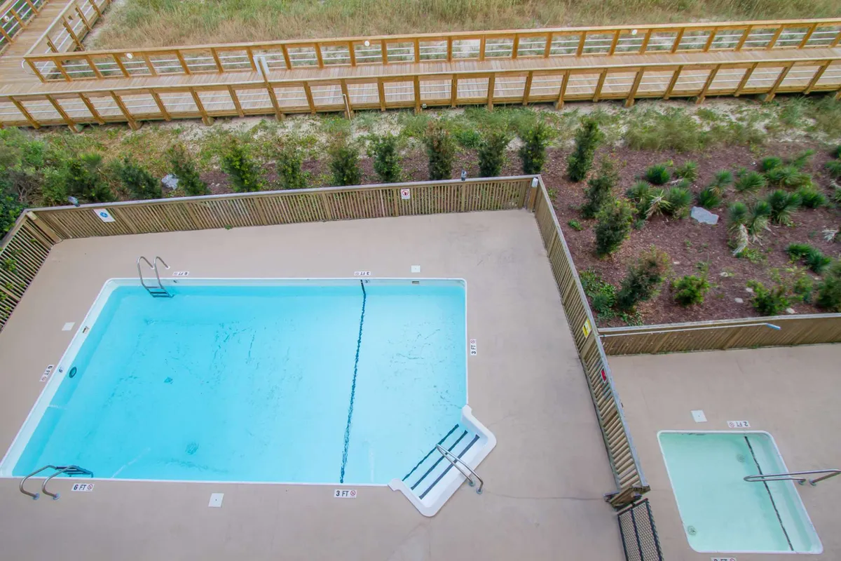 A sparkling community swimming pool surrounded by deck chairs and lush greenery. There's an inviting atmosphere, perfect for families to relax, cool off, and play together after a day at the beach.