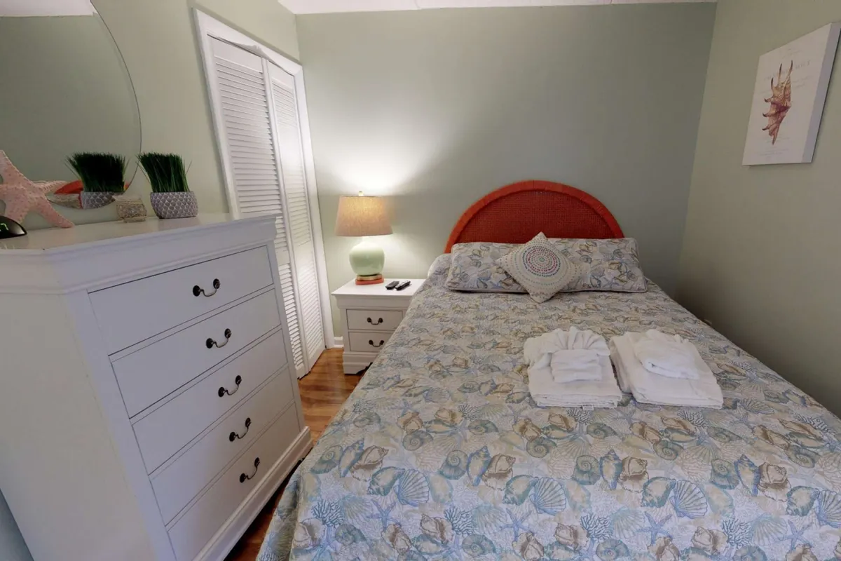 A tranquil bedroom featuring a neatly made bed with decorative pillows, a dressing table with a mirror, and a nightstand with a softly glowing lamp. Soft natural light filters in, and the room exudes a fresh, peaceful atmosphere ideal for relaxation.