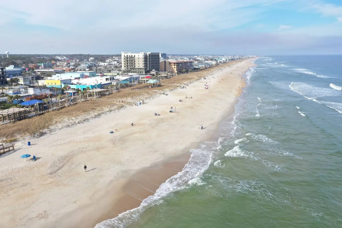 A vibrant scene of the beach, ocean, boardwalk, nearby buildings, and groups of people enjoying various activities such as sunbathing, walking, swimming, and socializing. The atmosphere is bustling and lively, suggesting a communal setting ideal for groups.
