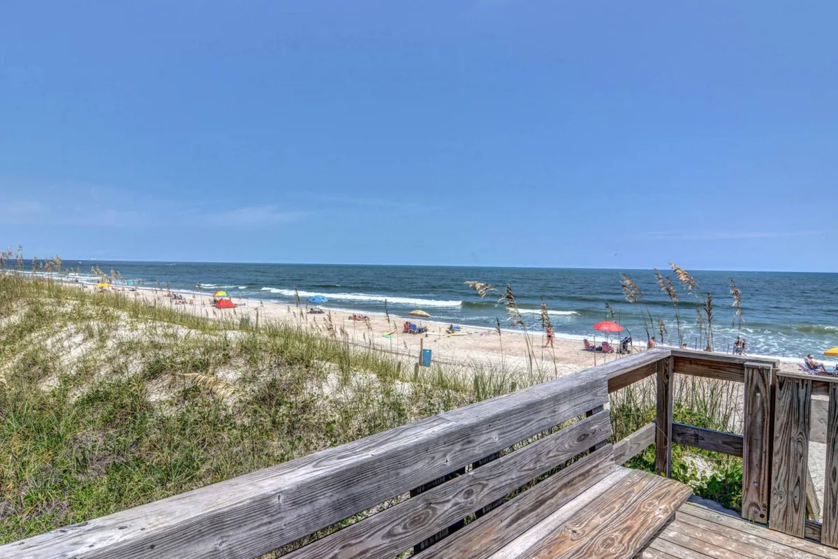 A cheerful beachfront scene featuring colorful umbrellas dotting the sand, families relaxing and playing, and a wide boardwalk that leads directly to the ocean. The sky is bright, and the atmosphere is bustling with summer fun.
