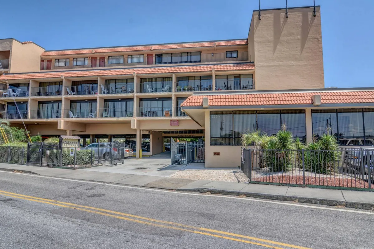 This image presents the exterior of the condo building, highlighting multiple balconies with seating, potted plants, and the covered parking area below. The building's facade is welcoming, and the parking area is clearly visible, emphasizing ease of access for residents.