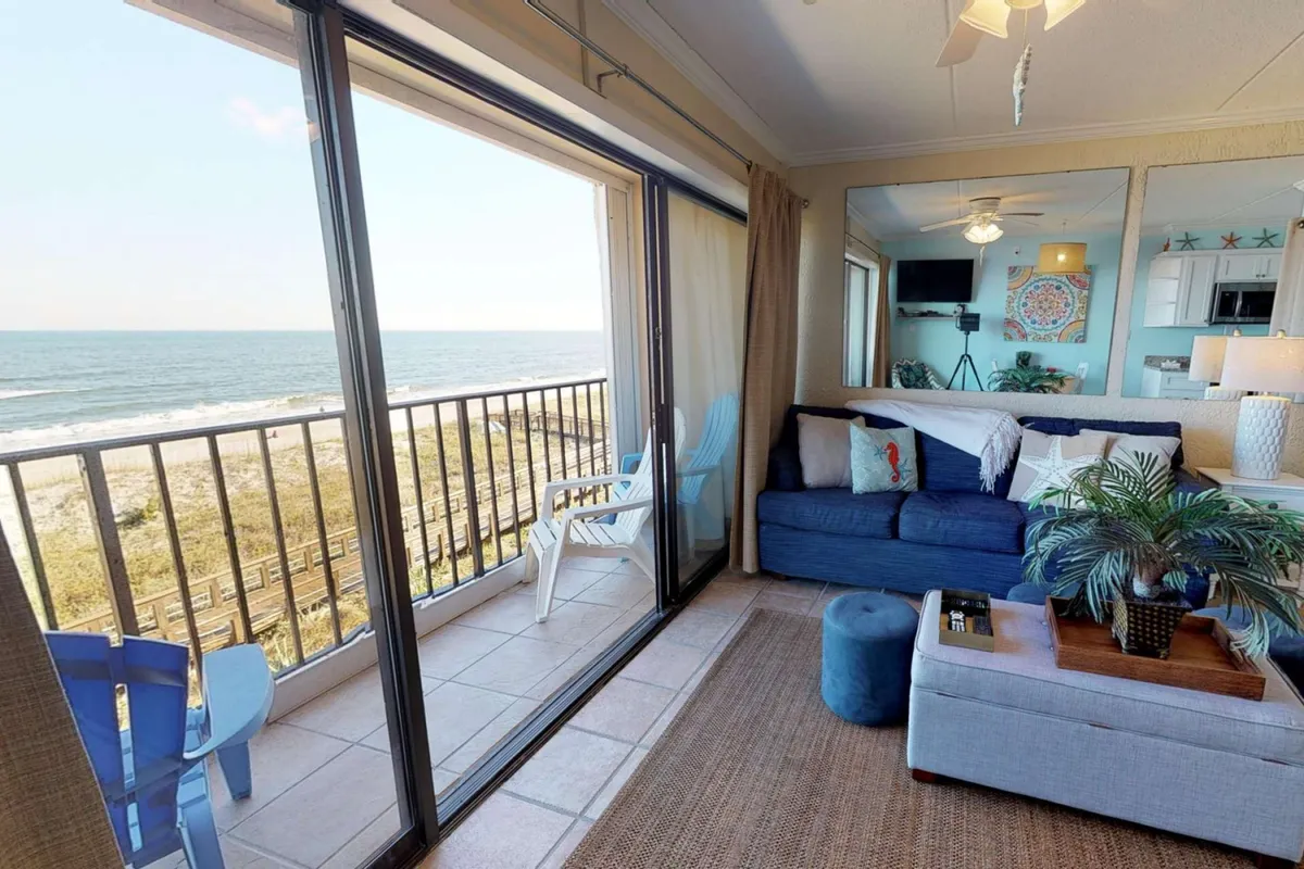 A serene living room with a blue couch, large glass doors opening to a private balcony, and an outdoor seating area overlooking the ocean. The image captures the transition from indoor comfort to outdoor relaxation.