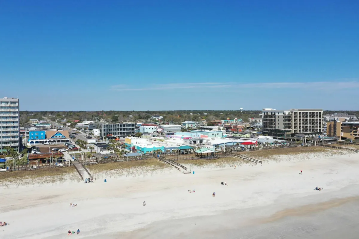 A lively view of the beach with colorful buildings along the boardwalk, people strolling, sunbathing, and enjoying local attractions. The boardwalk is evident, and the environment is vibrant and full of activity, suggesting proximity to shops, entertainment, and eateries.