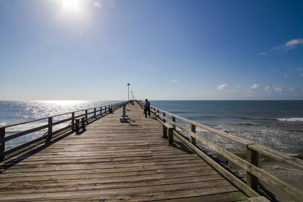 A picturesque exterior view of a wooden pier stretching over the ocean, illuminated by classic lamp posts. People stroll along the pier, immersed in the tranquil surroundings, with the sun rising over the water in the background, setting a serene, romantic mood.