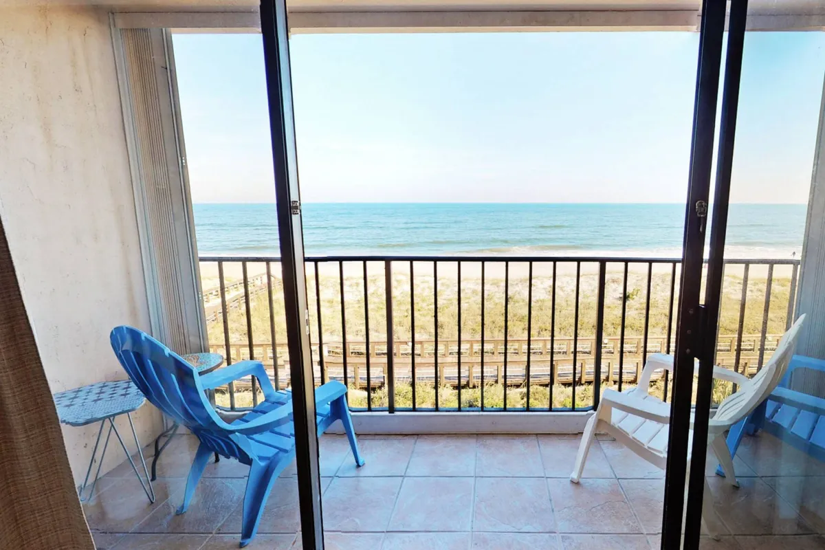 The image features a stunning oceanfront balcony at the condo, with inviting blue and white chairs positioned to capture sweeping views of the Atlantic. An open sliding door connects the bright interior to the tranquil outdoor space, while soft sunlight bathes the scene. The vantage point promises a front-row seat to sunrises, salty breezes, and relaxing moments with loved ones.